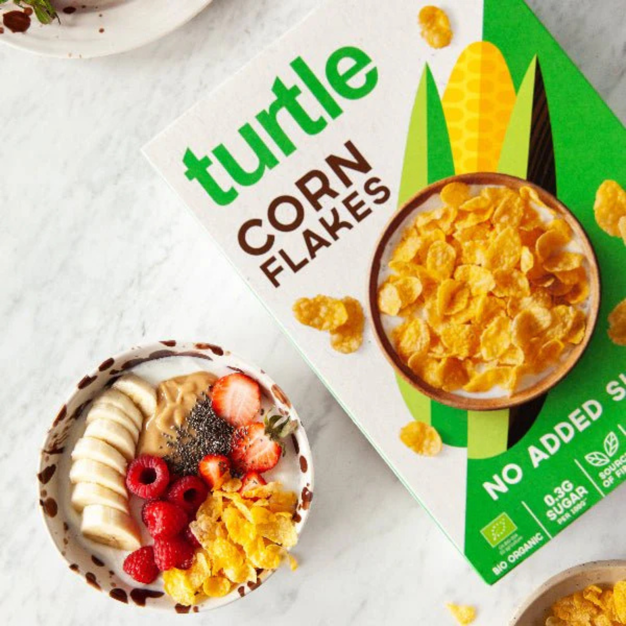 Top view of Turtle organic cornflakes box next to a breakfast bowl with gluten-free flakes, banana, raspberries, strawberries, chia seeds, and peanut butter